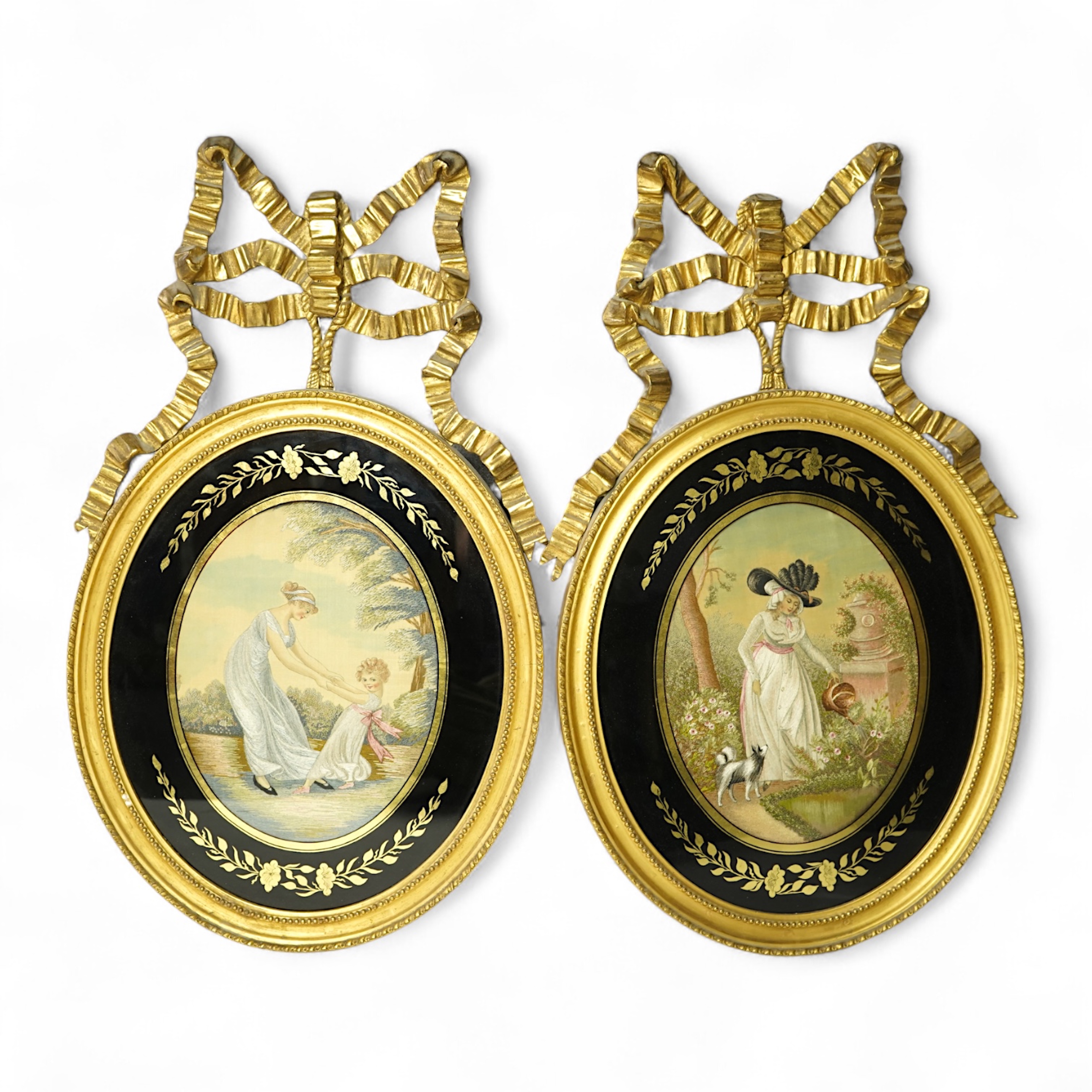 A pair of early 20th century oval silk embroideries, one of a mother and child dancing in a garden, the other of a lady in a large brimmed hat, watering flowers in by a classical stone urn, watched by her dog. Framed in